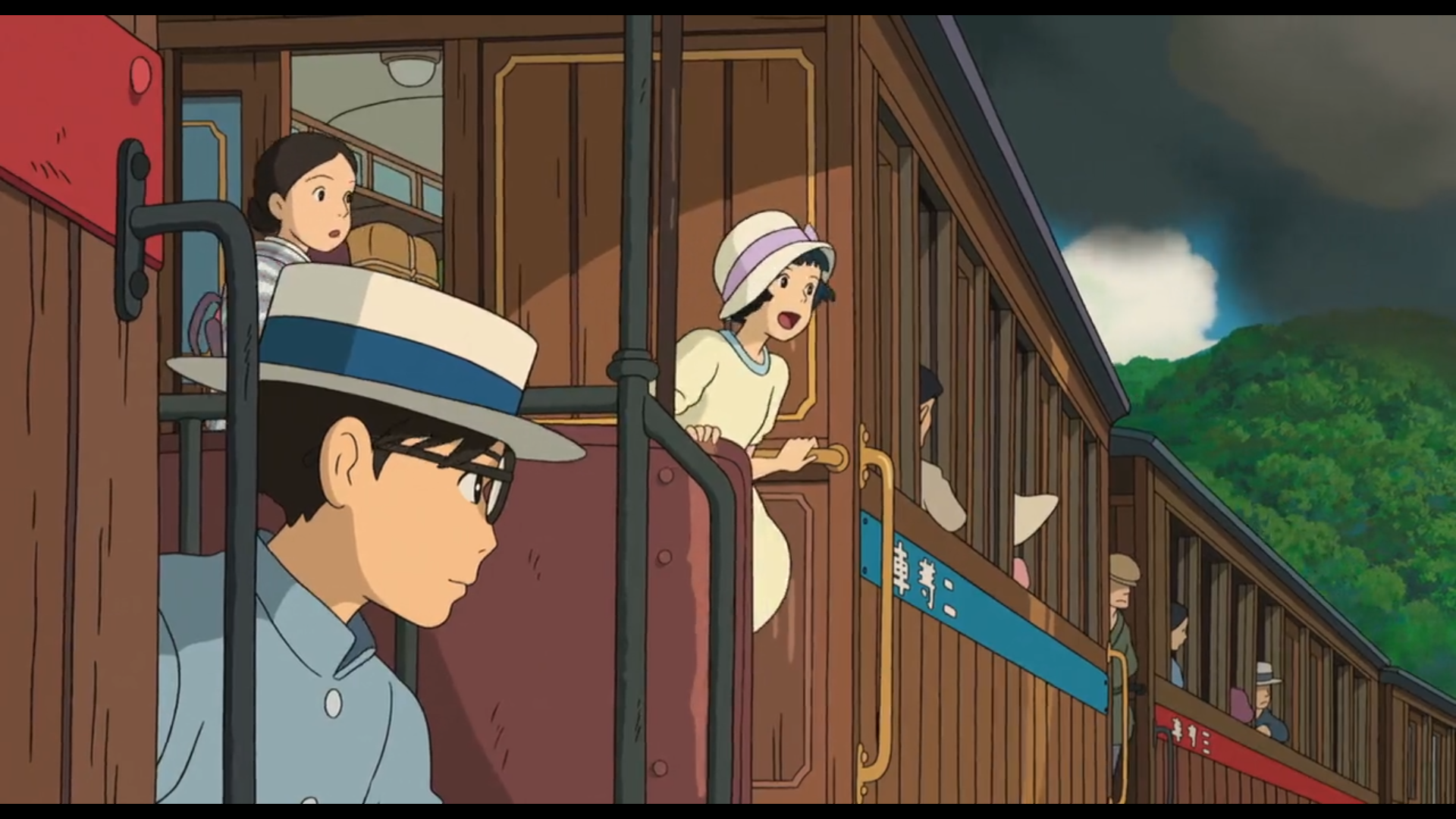 Scène du film Le Vent se lève
Au premier plan, Jiro, jeune homme japonais, aux cheveux noirs, yeux noirs, lunettes rondes, portant un habit bleu clair et un chapeau blanc avec un ruban bleu foncé, assis sur la une plateforme entre deux compartiments, il a le regard vers la personne à sa gauche.
Derrière justement, une jeune fille japonaise (on apprendra plus tard dans le film que son nom est Nahoko), aux cheveux noirs, yeux noirs, portant une robe claire et un chapeau avec un ruban mauve, se tient debout au bord de la plateforme, l'air émerveillé. Une femme un peu plus âgée, les cheveux foncés et le yeux noirs, portant un kimono rayé bleu et blanc,  la regarde avec appréhension.
Les wagons sont de couleur bois avec des bandes sous les fenêtres qui sont successivement rouge, bleue, puis à nouveau rouge, à chaque changement de compartiment.
On aperçoit diverses personnes aux fenêtres des wagons.
On aperçoit en arrière plan des arbres verts et la fumée du train cachant le ciel.