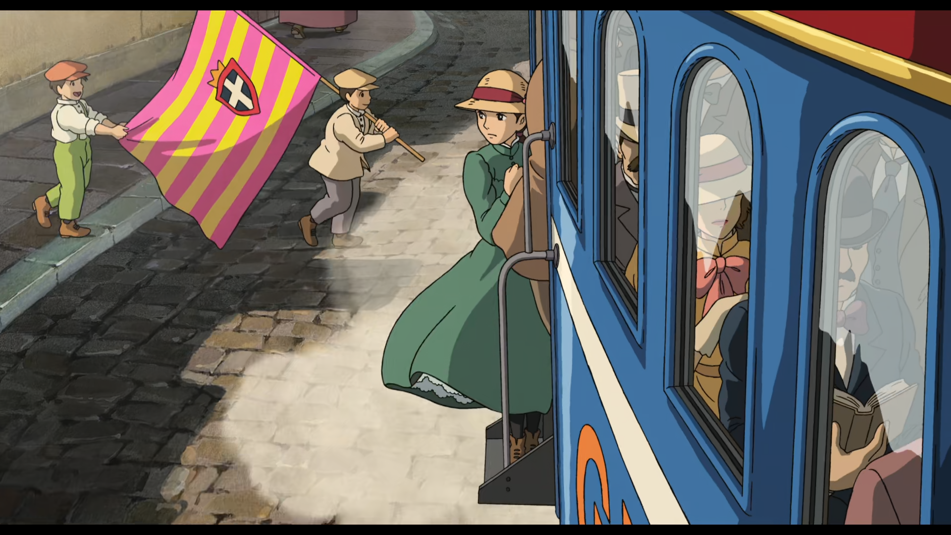 Scène du Chateau Ambulant
Sophie, jeune femme de type européen d'une vingtaine d'années, vêtue d'une longue robe verte et portant un chapeau en oseille clair avec un ruban bordeaux cachant ses cheveux, agrippée au wagon d'un tram, se tenant sur le marche-pied. Elle porte le regard vers la gauche à l'opposé du tram sur la droite.
Sur la droite, la rame de tram, bleue avec une bande blanche à mi hauteur et un toit rouge bordé de dorures, à travers les fenêtres on distingue que beaucoup de monde se tient dedans, entre autre, un homme moustachu, habillé en noir plongé dans la lecture d'un ouvrage à la couverture marron, une dame derrière, avec un ruban rose autour du cou et un chapeau lui cachant les yeux, et derrière un homme avec un haut de forme blanc. Dépassant de la porte du tram, on aperçoit les habits marrons d'un passager pas tout à fait à l'intérieur.
Sur la gauche de l'image, deux garçons traversent la rue pavée, de la gauche vers la droite. Le premier porte une veste et un béret de couleur claire, un pantalon gris et des chaussures marrons, et tient de ses deux mains le manche d'un drapeau rayé rose et jaune avec un blason contenant une croix blanche, sur un fond bleu foncé aux pourtours rouges surmonté d'un couronne, trainant derrière l'enfant. Le deuxième garçon porte une chemise blanche, un béret orange, un pantalon vert pistache et des souliers clairs, et tient le drapeau par son extrémité avec une mine enjouée. 