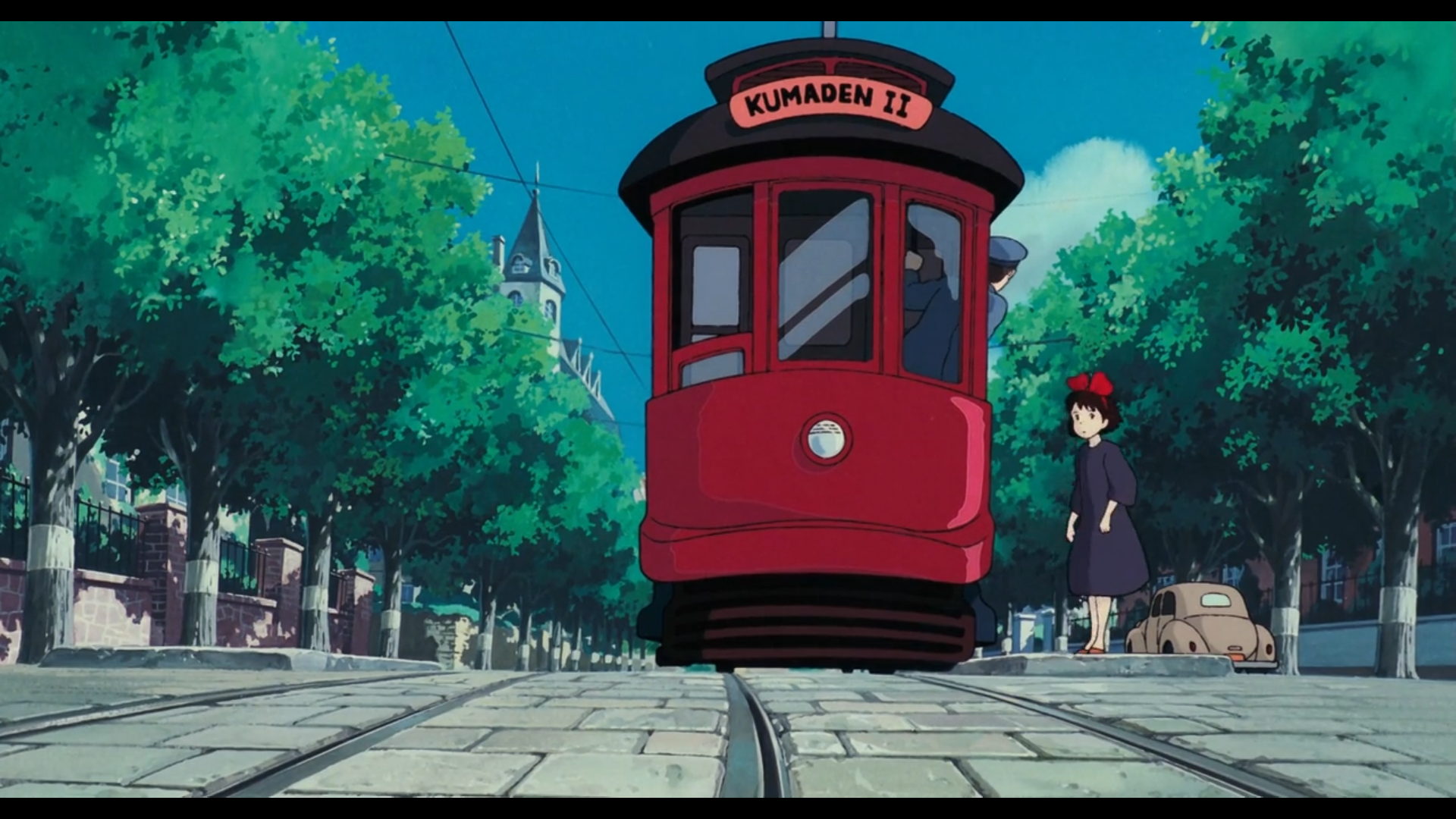 Scène de Kiki la petite sorcière
Plan d'ensemble où Kiki, jeune fille de type européen, aux cheveux noirs, yeux noirs, portant une robe sombre et un nœud rouge dans les cheveux, se tient à côté d'un tram rouge avec un toit noir et un panneau en haut sur lequel est inscrit "Kumaden II". La tête d'un homme aux cheveux noirs portant un costume bleu et une casquette bleue qui semble être le conducteur, dépasse d'une des fenêtres du wagon.
Ils sont dans une rue pavée, bordée d'une rangée d'arbres de part et d'autre, cachant en partie des murets en brique surmontés de grilles, indiquant qu'il s'agit d'un quartier résidentiel.
On aperçoit aussi une tour dépassant des arbres, et sur la droite, garée sur le côté, une voiture décapotable de couleur claire dont le style rappelle les années 50.
