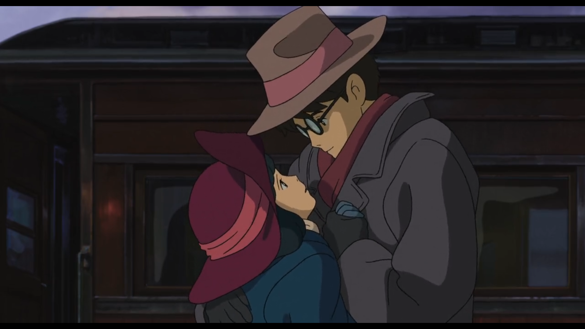 Scène du film Le Vent se lève
Jiro, homme japonais, cheveux noirs, yeux noirs, lunettes rondes, emmitouflé dans un manteau gris foncé avec une écharpe bordeaux autour du cou, un chapeau marron avec un ruban rose sur la tête et des gants noirs, serre fort contre lui Nahoko, femme japonaise, cheveux noirs, yeux noirs, portant un manteau bleu foncé, un chapeau bordeaux avec un ruban rose et des gants bleus, en lui tenant les mains. Ils échangent un regard rempli de tendresse.
En arrière plan, on distingue la forme d'un compartiment de train, de couleur sombre, indiquant que la scène se déroule dans une gare.